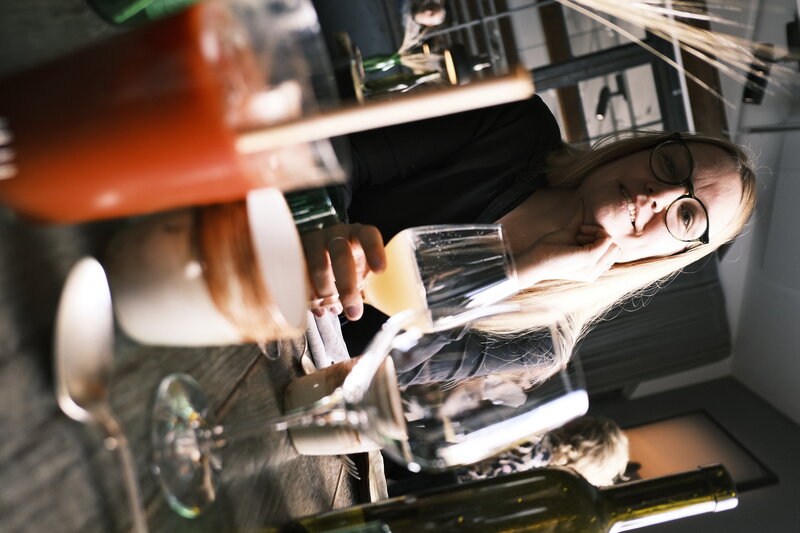 Woman with glasses sitting at a restaurant table holding a glass of white wine, drinks and small dishes in the foreground