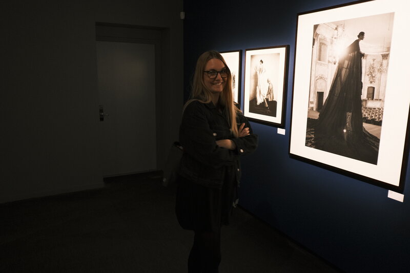 Woman smiling with arms crossed in a dark gallery room, large black-and-white exhibition prints on the dark blue wall behind her