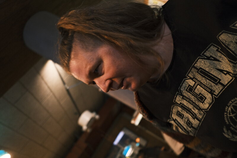 Close portrait of a person with one side of their head shaved and long hair on the other, looking sideways in warm bar light