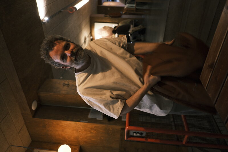 Bearded man in a white t-shirt leaning over a table and grinning toward the camera
