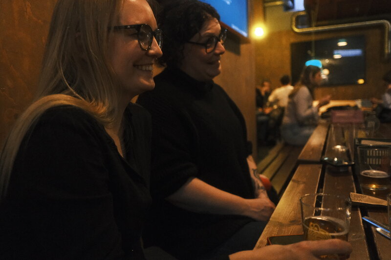 Two women with glasses laughing together at a long bar counter in warm, dim lighting