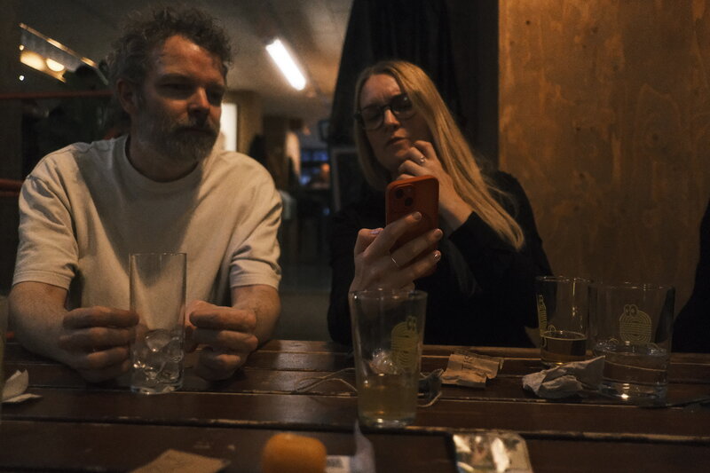 Man and woman at a bar table late in the evening — he holds an empty glass, she looks at her phone