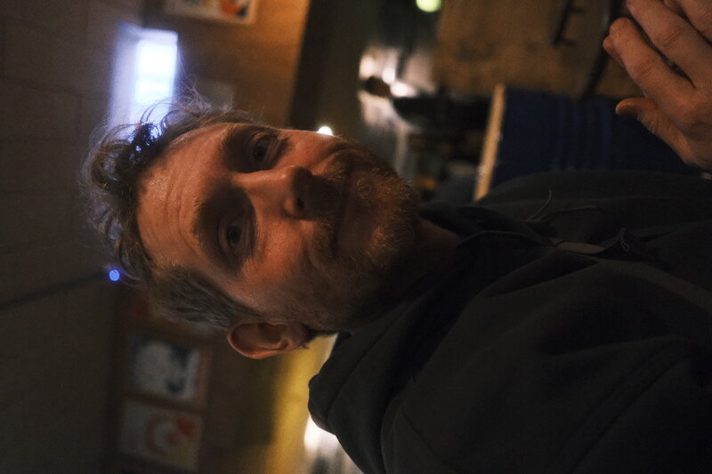 Close portrait of a man in a dark hoodie looking toward the camera in a warm-lit bar